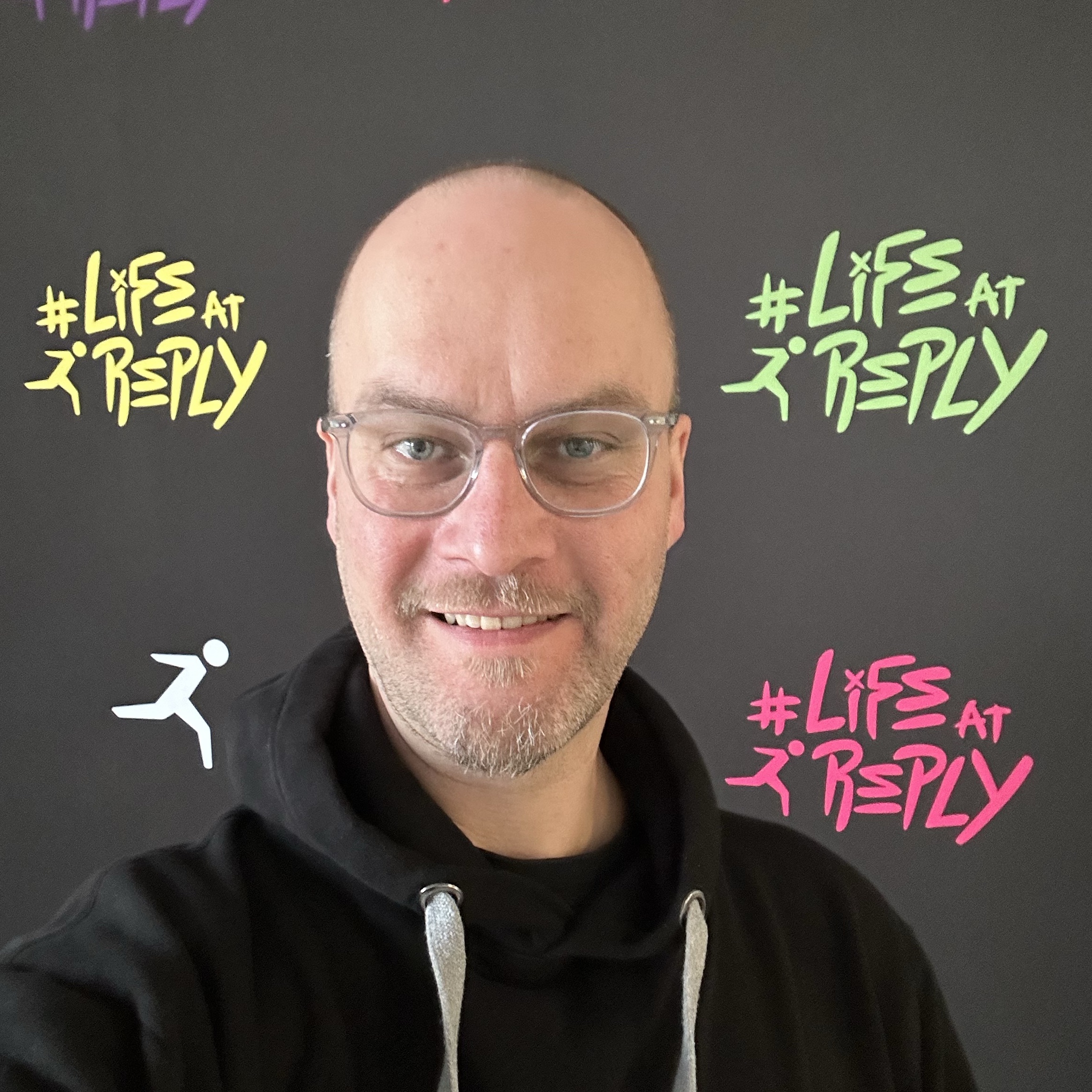 A smiling man with glasses poses in front of a black backdrop featuring colorful text and a running figure, promoting '#Life at Reply.'